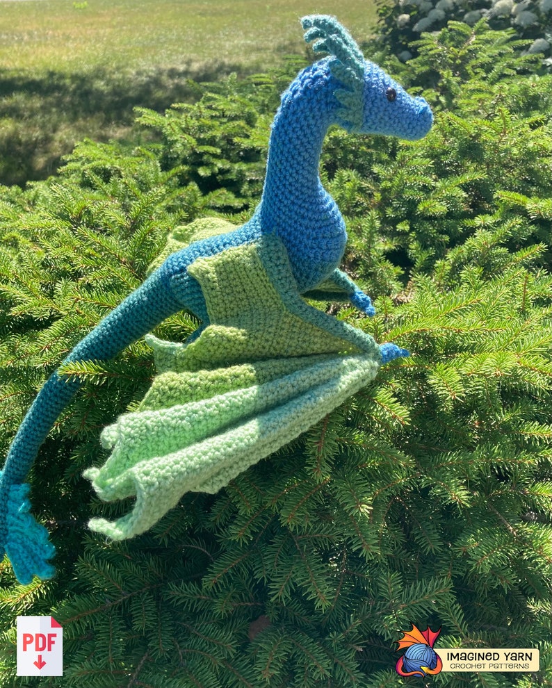 Peut inclure: Un dragon bleu et vert en crochet avec les ailes d&eacute;ploy&eacute;es. Le dragon est assis sur un lit de branches de pin vertes.