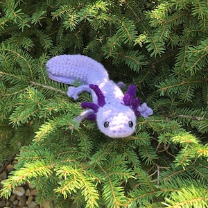 May include: A crocheted axolotl plush toy in shades of purple and lavender, with large, black eyes. The toy is sitting on a bed of green pine needles.