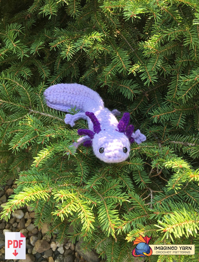 May include: A crocheted axolotl plush toy in shades of purple and lavender, with large, black eyes. The toy is sitting on a bed of green pine needles.