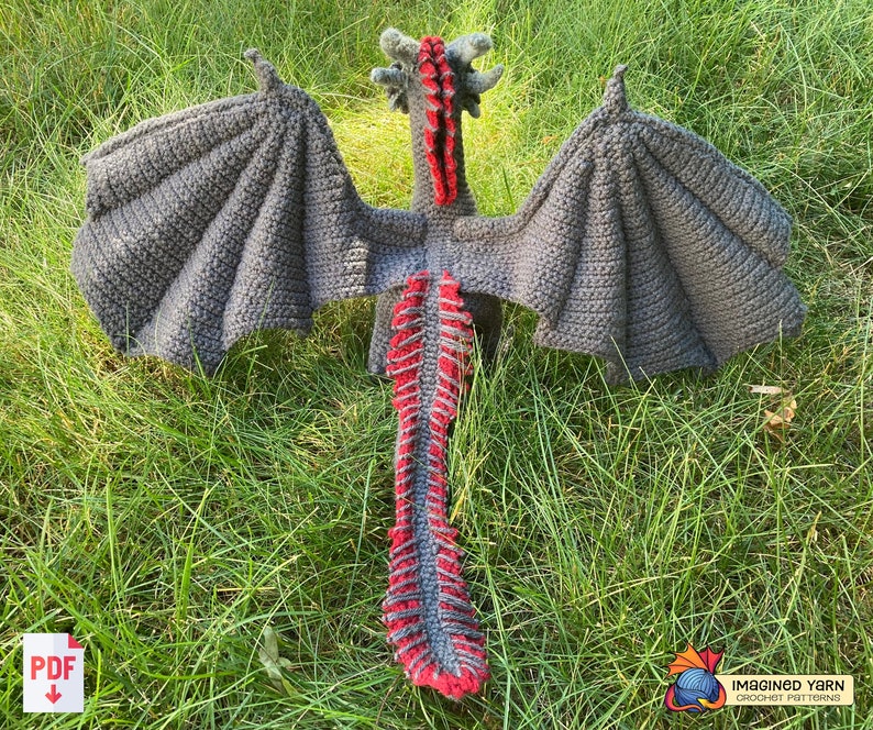 Peut inclure: Un jouet de dragon en crochet avec des ailes grises et une queue rouge et grise. Le dragon est couch&eacute; sur le dos, les ailes d&eacute;ploy&eacute;es.