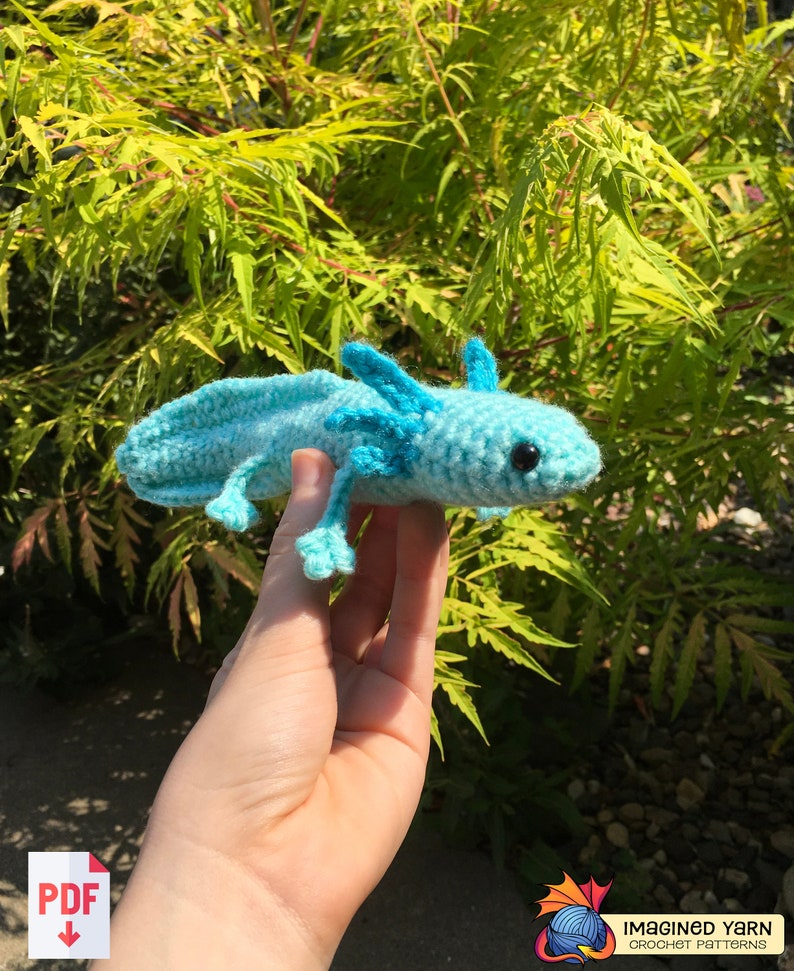 May include: A crocheted blue axolotl plush toy with black eyes. The toy is being held in a person's hand. The background is a green leafy bush.