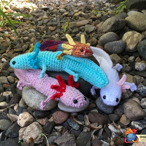 May include: Four crocheted axolotl plushies in different colors: light blue, pink, white, and orange with yellow accents. The plushies are sitting on a bed of small gray rocks.