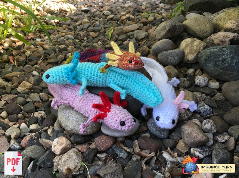 May include: Four crocheted axolotl plushies in different colors: light blue, pink, white, and orange with yellow accents. The plushies are sitting on a bed of small gray rocks.