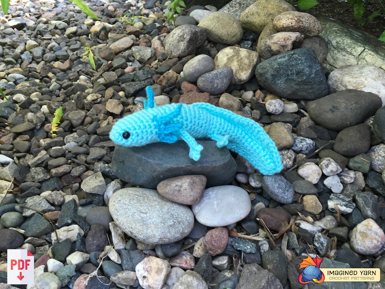 May include: A crocheted blue axolotl, a type of salamander, is sitting on a rock. The axolotl is a popular pet and is known for its unique appearance.