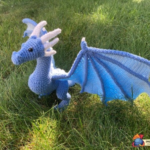 Peut inclure: Un dragon en crochet bleu clair avec des cornes et des ailes blanches. Le dragon est en position avec ses ailes d&eacute;ploy&eacute;es et est assis sur un lit d'herbe verte.