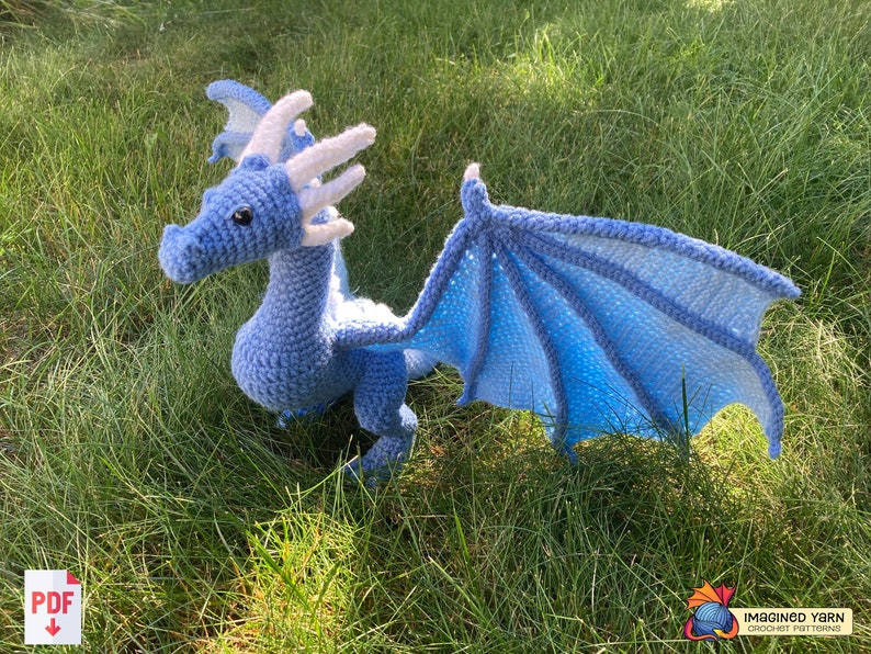 Peut inclure: Un dragon en crochet bleu clair avec des cornes et des ailes blanches. Le dragon est en position avec ses ailes d&eacute;ploy&eacute;es et est assis sur un lit d'herbe verte.
