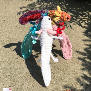 May include: Three crocheted axolotl plushies. One is white, one is rainbow, and one is orange and yellow. The plushies are standing on a sandy surface.