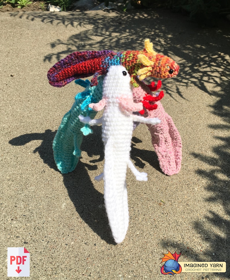 May include: Three crocheted axolotl plushies. One is white, one is rainbow, and one is orange and yellow. The plushies are standing on a sandy surface.