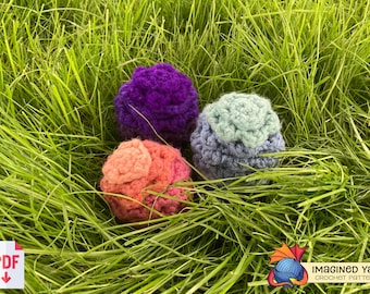 May include: Three crocheted balls in different colors: purple, blue and orange. The balls are sitting on a bed of green grass.