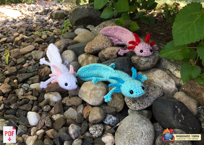 May include: Three crocheted axolotl plushies in different colors: white, pink, and blue. The plushies are sitting on a bed of rocks.