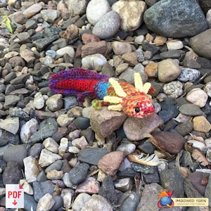 May include: A crocheted axolotl toy with a red, yellow, blue, and purple color scheme. The toy is sitting on a bed of small rocks.