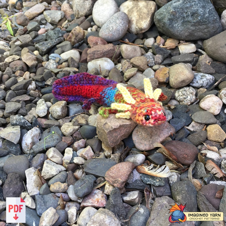 May include: A crocheted axolotl toy with a red, yellow, blue, and purple color scheme. The toy is sitting on a bed of small rocks.