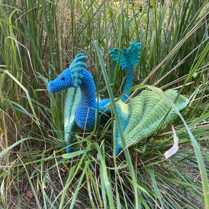Peut inclure: Un dragon bleu et vert en crochet avec des ailes, assis dans de hautes herbes vertes.
