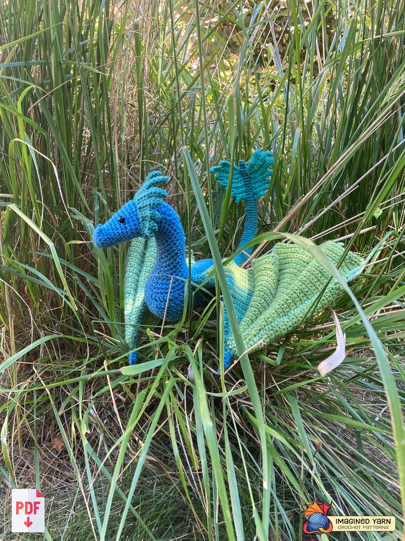 Peut inclure: Un dragon bleu et vert en crochet avec des ailes, assis dans de hautes herbes vertes.