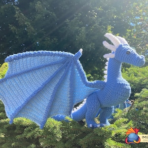 Peut inclure: Un dragon bleu en crochet avec des cornes et des ailes blanches. Le dragon est en position debout sur un lit d'aiguilles de pin vertes.