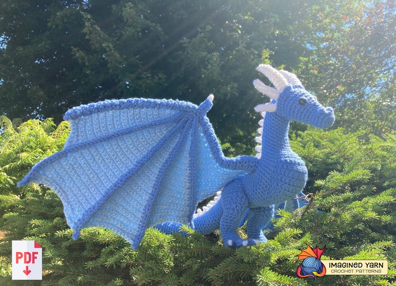 Peut inclure: Un dragon bleu en crochet avec des cornes et des ailes blanches. Le dragon est en position debout sur un lit d'aiguilles de pin vertes.