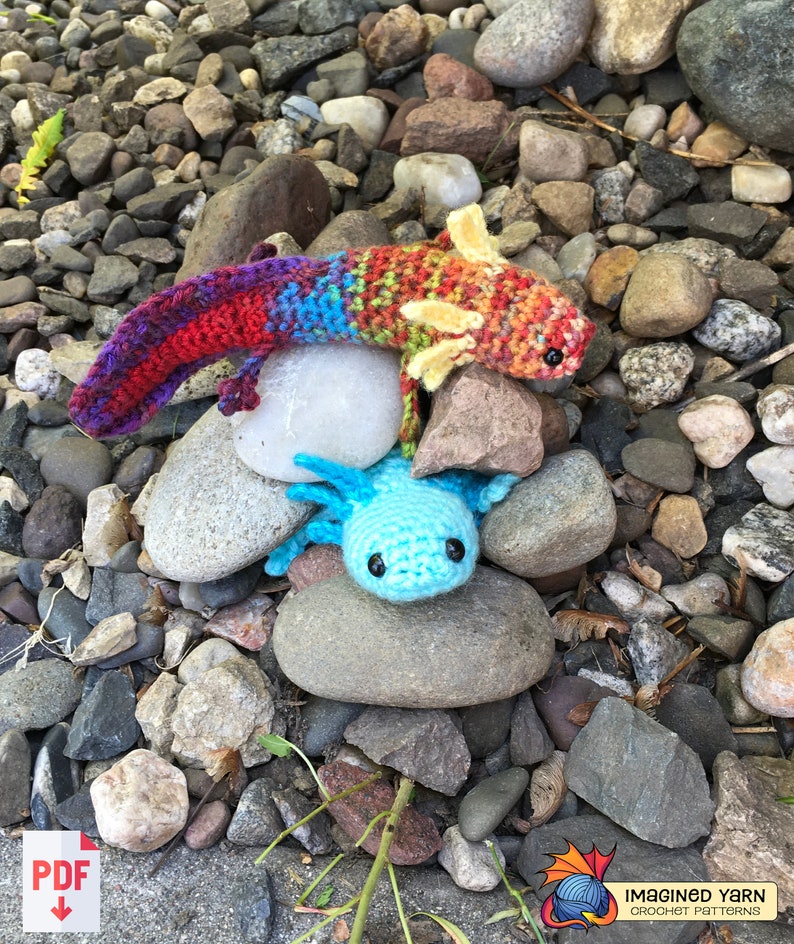 May include: Two crocheted axolotl plushies, one is rainbow colored and the other is blue, sitting on a bed of rocks.
