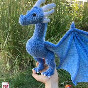 Peut inclure: Un dragon bleu crochet&eacute; avec des cornes et des ailes blanches. Le dragon est debout sur la main d'une personne.