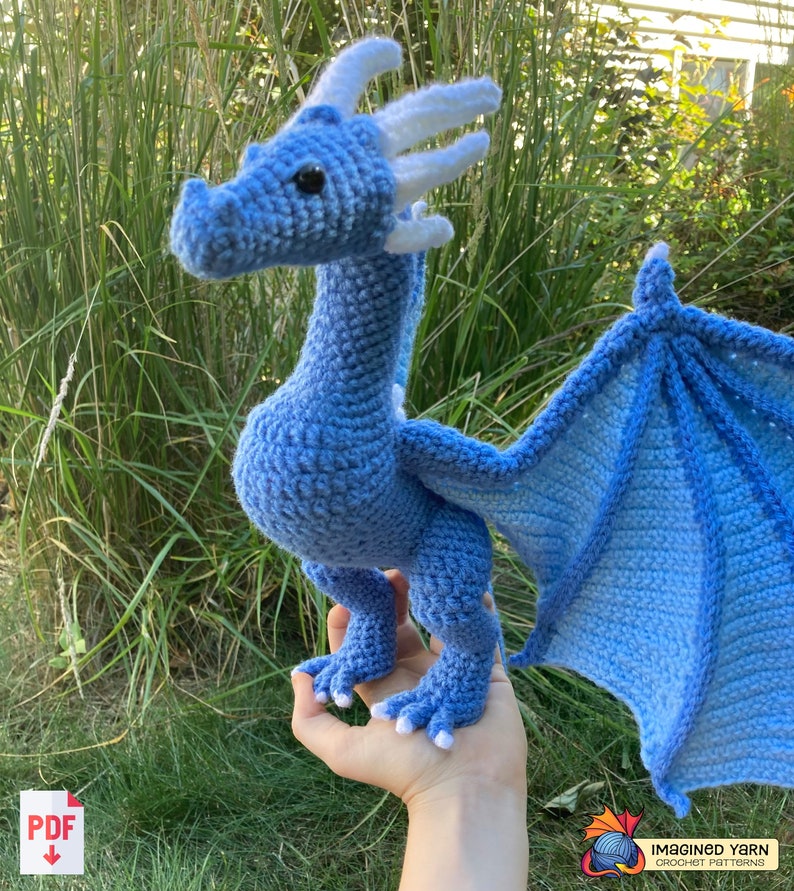 Peut inclure: Un dragon bleu crochet&eacute; avec des cornes et des ailes blanches. Le dragon est debout sur la main d'une personne.