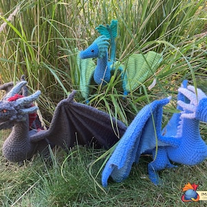 Peut inclure: Trois peluches de dragon au crochet de diff&eacute;rentes couleurs&nbsp;: gris, bleu et vert. Les dragons sont pos&eacute;s dans une zone herbeuse, les ailes d&eacute;ploy&eacute;es.