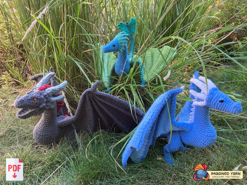 Peut inclure: Trois peluches de dragon au crochet de diff&eacute;rentes couleurs&nbsp;: gris, bleu et vert. Les dragons sont pos&eacute;s dans une zone herbeuse, les ailes d&eacute;ploy&eacute;es.