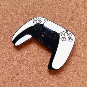 May include: A black and white PlayStation 5 controller pin. The pin features the iconic design of the controller, with white sides and black center. The pin has a detailed design of the buttons and directional pad.