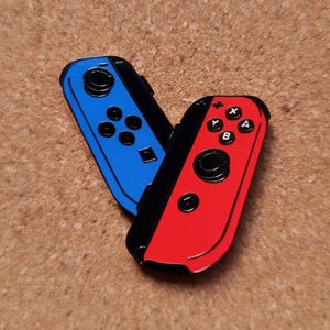May include: A pair of blue and red video game controller pins. The blue controller has black buttons and a black border. The red controller has black buttons and a black border. The controllers are angled towards each other.