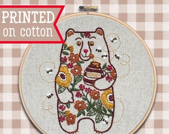 Pode incluir: Arte de bordado em forma de aro que representa um urso marrom segurando um pote de mel, cercado por flores e abelhas. O texto "Honeybear" e "Option A on Natural" está impresso abaixo do aro.