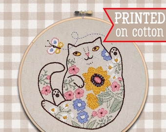 Pode incluir: Desenho bordado de um gato branco com olhos amarelos, rodeado de flores coloridas, sobre um fundo de tecido bege. O desenho está em um aro circular. O texto "Playtime Kitty" e "Kindred Spirit ART STUDIO" também estão incluídos.