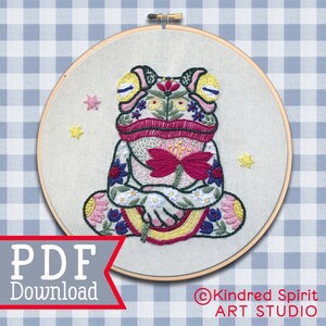 May include: Embroidered hoop art featuring a colorful frog with floral designs and a rainbow. The frog is sitting in a meditative pose with its eyes closed. The hoop is surrounded by a blue and white gingham background. The text "PDF Download" and "Kindred Spirit ART STUDIO" are visible in the image.