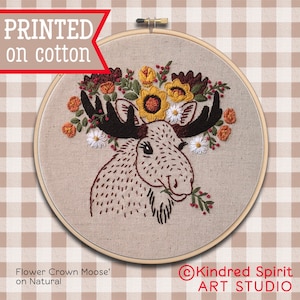 Op de afbeelding: Een geborduurde eland met een bloemenkrans op een natuurlijke stoffen achtergrond. De borduurwerk is in tinten bruin, geel en rood. De tekst "Flower Crown Moose" en "Kindred Spirit Art Studio" is onder de afbeelding gedrukt.