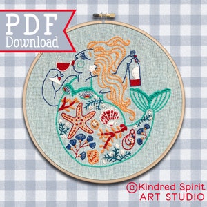 May include: Embroidered hoop art featuring a mermaid with a glass of wine and a bottle of wine. The mermaid is surrounded by sea life, including starfish, seashells, and coral. The hoop is light blue with a wooden frame.