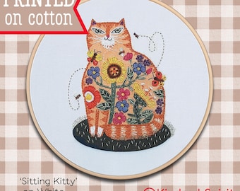 Pode incluir: Desenho bordado de um gato laranja sentado em um fundo preto, cercado por flores coloridas e abelhas. O desenho é impresso em tecido de algodão branco e emoldurado em um aro de madeira. O texto 'Sitting Kitty' e 'on White' é impresso abaixo do aro. O texto 'Kindred Spirit ART STUDIO' é impresso no canto inferior direito.