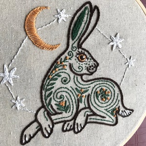 May include: Embroidered hoop art featuring a green rabbit with a crescent moon and stars in the background. The rabbit is surrounded by intricate floral designs and has a brown eye.