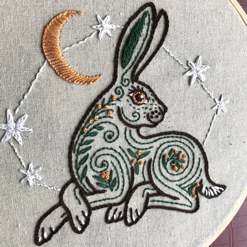 May include: Embroidered hoop art featuring a green rabbit with a crescent moon and stars in the background. The rabbit is surrounded by intricate floral designs and has a brown eye.
