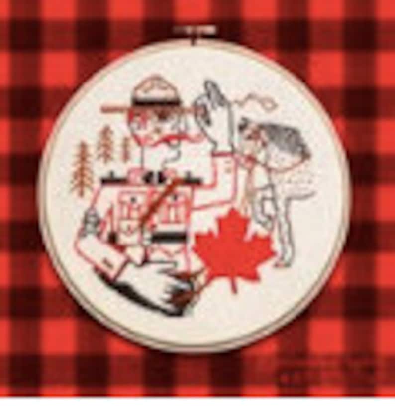 May include: Embroidered hoop art featuring a cartoon illustration of a Canadian Mountie in uniform with a red maple leaf and a wolf beside him. The background is a red and black plaid pattern.