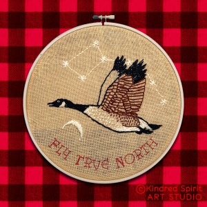 May include: Embroidered hoop art featuring a stylised goose flying with a constellation of stars above it. The text "Fly True North" is stitched in red thread on a beige background.
