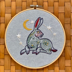 May include: Embroidered hoop art featuring a stylized green and red rabbit with a yellow crescent moon and white stars on a light blue background.