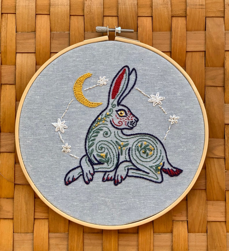 May include: Embroidered hoop art featuring a stylized green and red rabbit with a yellow crescent moon and white stars on a light blue background.