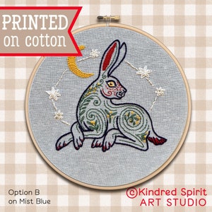 May include: Embroidered hoop art featuring a stylized grey rabbit with red, yellow, and green details. The rabbit is surrounded by a crescent moon and stars on a light blue fabric background. The text "Kindred Spirit ART STUDIO" is visible in the lower right corner.