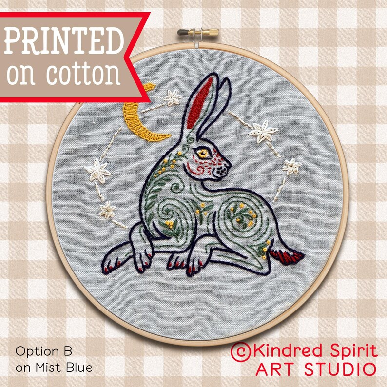 May include: Embroidered hoop art featuring a stylized grey rabbit with red, yellow, and green details. The rabbit is surrounded by a crescent moon and stars on a light blue fabric background. The text "Kindred Spirit ART STUDIO" is visible in the lower right corner.