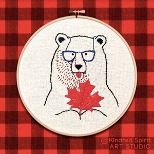 May include: Embroidered hoop art featuring a white polar bear wearing blue glasses and holding a red maple leaf. The background is a red and black checkered pattern. The text "Kindred Spirit ART STUDIO" is in the bottom right corner.