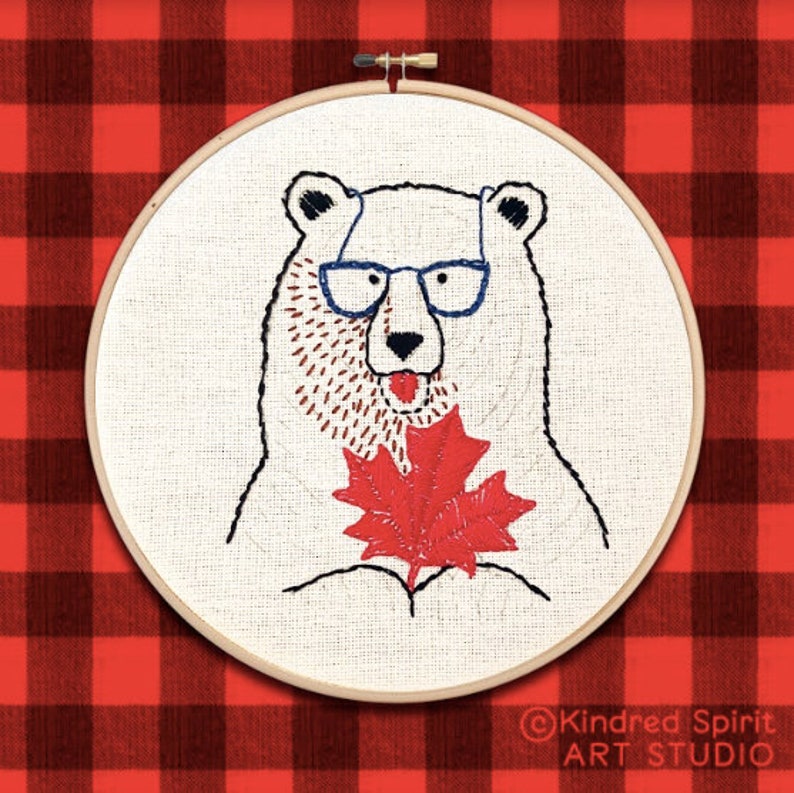 May include: Embroidered hoop art featuring a white polar bear wearing blue glasses and holding a red maple leaf. The background is a red and black checkered pattern. The text "Kindred Spirit ART STUDIO" is in the bottom right corner.