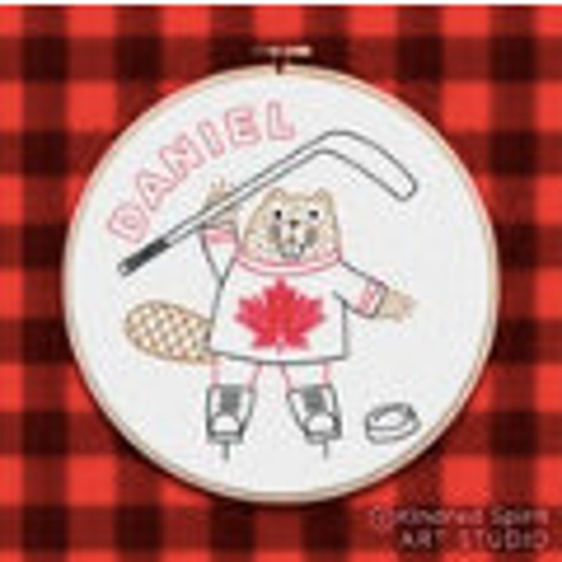 May include: Embroidered hoop art featuring a cartoon beaver wearing a red sweater with a Canadian flag on the front, holding a hockey stick and ice skates. The beaver's name, "Daniel", is written above the beaver in a white font.