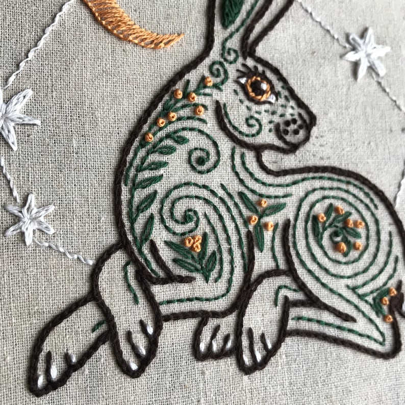 May include: Embroidered design of a green and brown hare with intricate details, including swirls and leaves. The hare is surrounded by a crescent moon and stars.