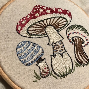 May include: Embroidered mushroom design on a beige fabric background. The mushrooms are red with white spots, blue and white striped, and green with red spots.