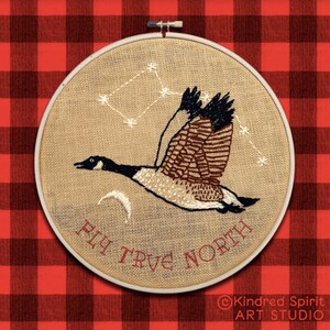 May include: Embroidered hoop art featuring a brown and white goose flying with a constellation of stars above it. The text "Fly True North" is stitched in red below the goose. The hoop is on a red and black checkered background.