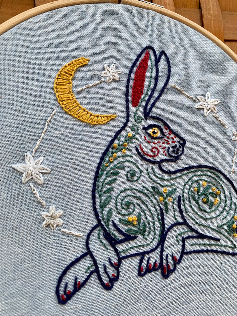 May include: Embroidered hoop art featuring a stylized blue rabbit with green and yellow floral details, a yellow crescent moon, and white stars on a light blue background.