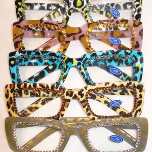 May include: An assortment of reading glasses with a leopard print design in various colors. Each pair features decorative rhinestones around the frames and lenses. The glasses have a +3.00 magnification.