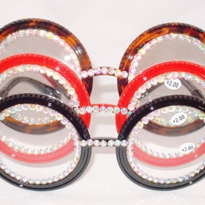 May include: Three pairs of round reading glasses with rhinestones around the lenses. The glasses are layered on top of each other, with the top pair being tortoise shell, the middle pair being red, and the bottom pair being black. Each pair has a +2.00 label.